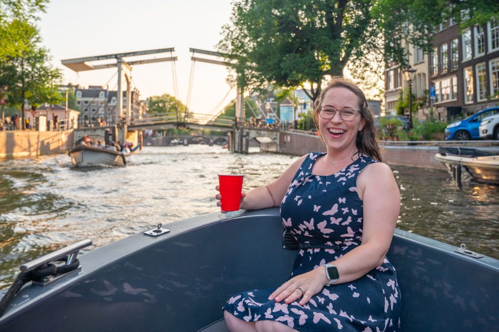 Woman on the front of a boat facing the camera smiling with a bridge behind her