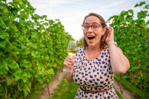 Woman in polka dot dress facing camera with glass of white wine in between vineyards