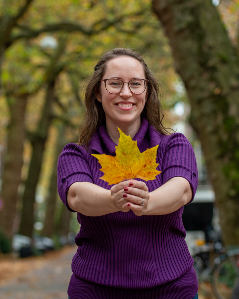 Woman in purple sweater holding one large green/yellow leaf towards the camera with a blurred background of trees in autumn colors